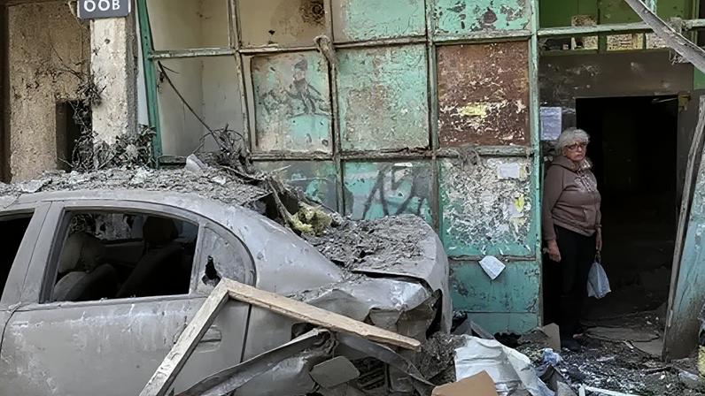 An elderly woman stands in the entrance to a damaged apartment block. To her left is a badly damaged car with no windows. Debris lies all around.