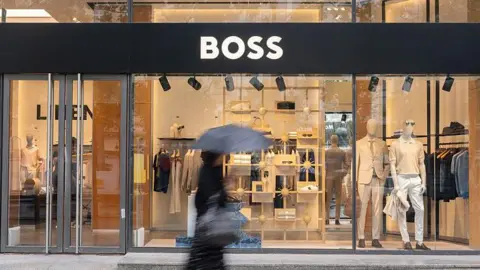 Getty Images The entrance of the Hugo Boss flagship fashion store in Paris, France. In the foreground, a blurred woman is walking past with an umbrella. Inside the shop, mannequins, shirts, shoes, and trousers are visible.