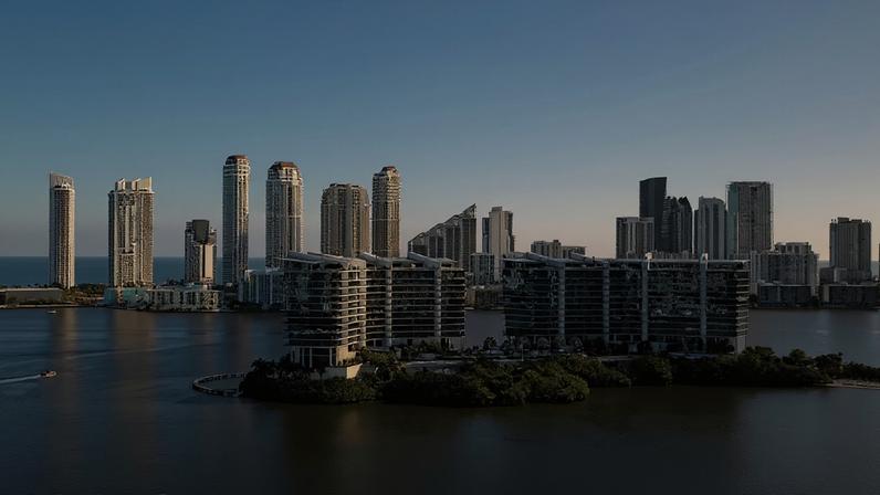 Vista aérea de uma cidade costeira com arranha-céus altos e modernos à beira-mar, sob um céu azul claro. Edifícios residenciais baixos e luxuosos ocupam uma ilha verde no centro da imagem, cercada por águas calmas. Um pequeno barco navega à esquerda.