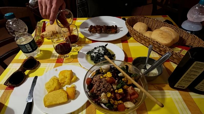 A table spread with salad, grilled meat skewers, polenta, greens, bread, and wine on a checkered tablecloth