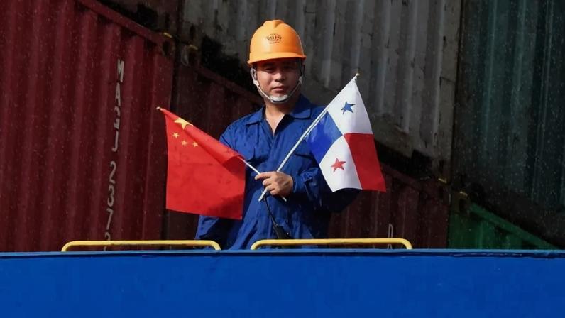 Um trabalhador vestido com macacão azul e capacete de proteção laranja segura as bandeiras da China e do Panamá