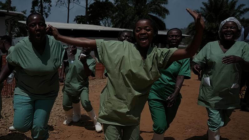 BBC Health workers wearing green scrubs and white wellie boots dance and sing on a dusty ground