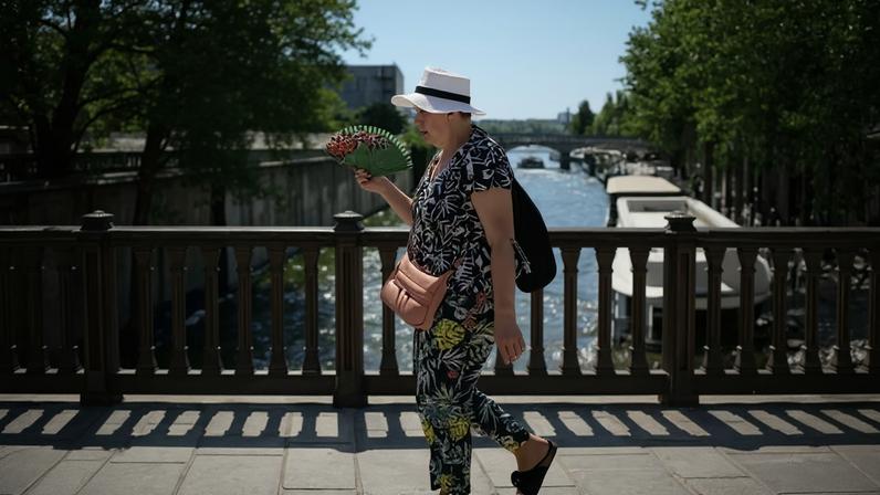 A woman in a sun hat uses a hand fan as she crosses a bridge over the Seine in Paris
