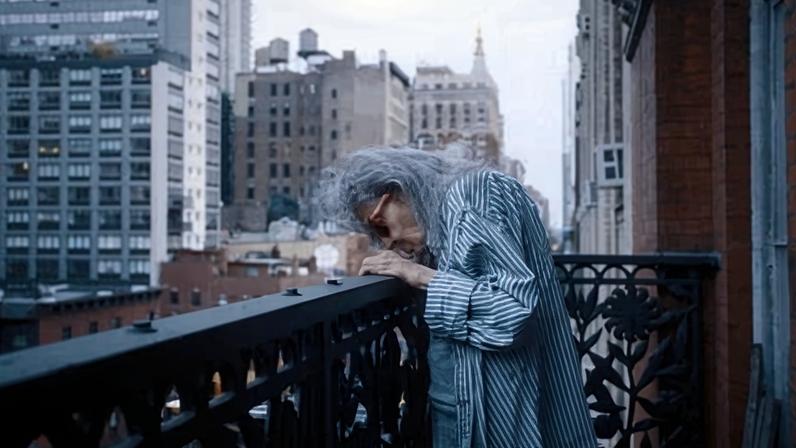 Bettina stands on a city balcony, head bowed and arms crossed