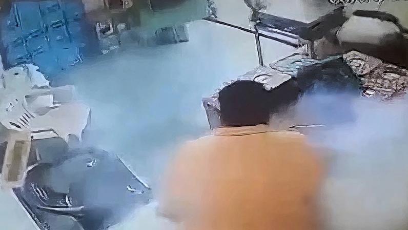 Registro de câmera de segurança dentro de um pequeno estabelecimento. Um homem de camisa laranja está próximo a um balcão, e há uma nuvem de fumaça ao redor.