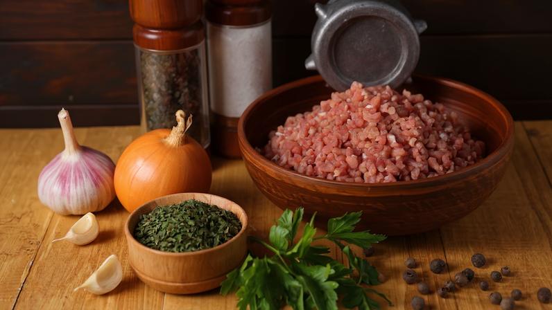 Moedor de carne manual com carne moída, especiarias e salsa sobre a mesa de madeira.