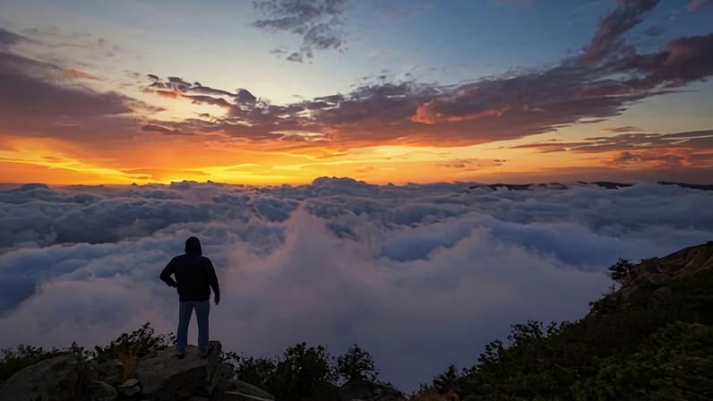 A man standing on a mountain during sunset, looking over low-lying clouds