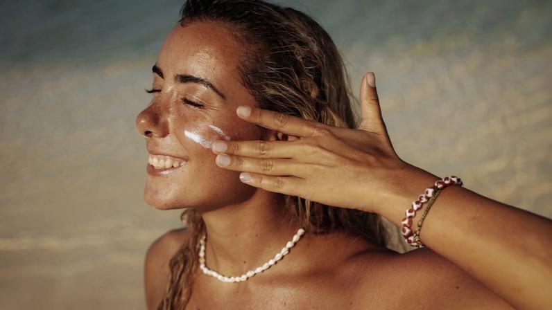 Foto colorida de mulher na praia e aplicando protetor solar no rosto - Metrópoles