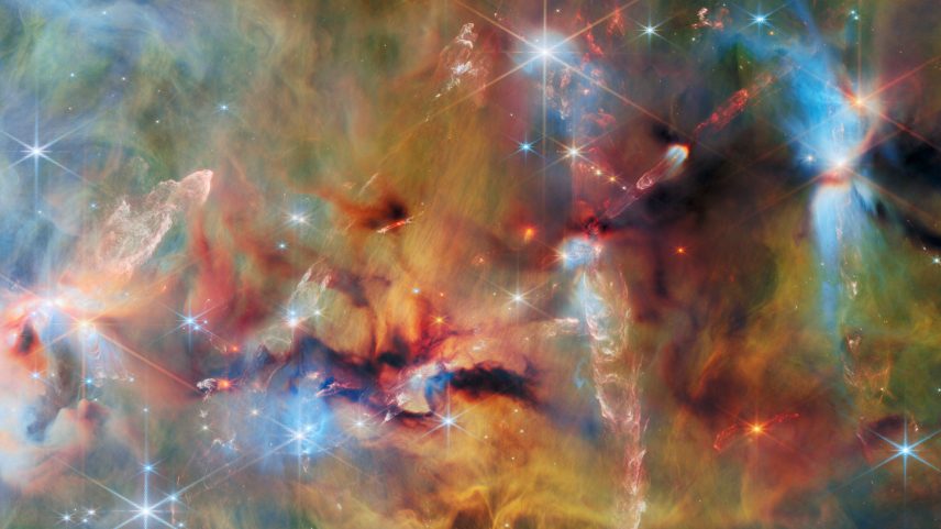 An area inside a star-forming molecular cloud. The background is covered with layers of gas and dust in blue, green and yellowish colors. Thicker clumps of cold dust, dark brown to black, block out light completely. Stars lie among and atop the clouds, from small orange ones to large white or blue ones. Waves and streams of glowing whitish gas are created by jets from protostars colliding with the surrounding material.