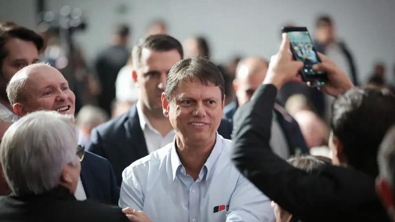 Tarcísio de Freitas, sorrindo e de braços cruzados, veste camisa social azul clara com distintivo no peito, em meio a uma multidão. Um homem careca ao fundo ri, e uma pessoa à direita filma com um celular.