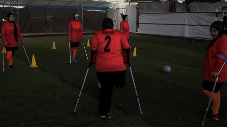 Mulheres atletas com deficiência física participando de um treino de futebol adaptado em um campo coberto de grama sintética.