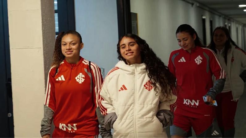 Quatro mulheres caminhando por um corredor interno, vestindo uniformes vermelhos e brancos do Sport Club Internacional, equipe de futebol de Porto Alegre.
