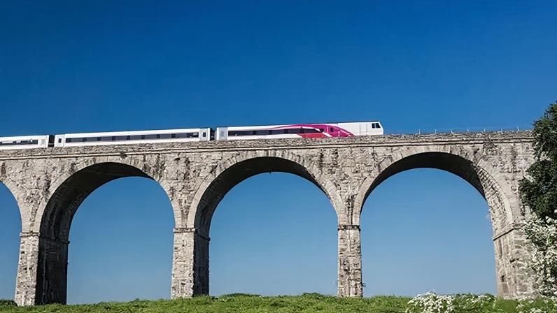 A bridge, on top of a field of grass, with a train crossing it. There is a blue sky in the background.