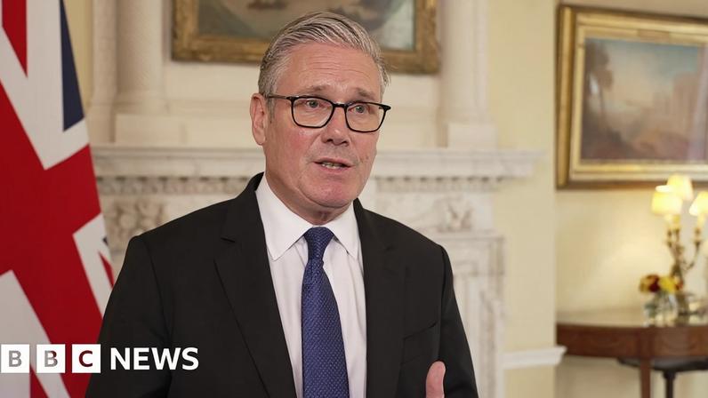 Sir Keir Starmer wears a black suit and blue tie standing in front of a British flag
