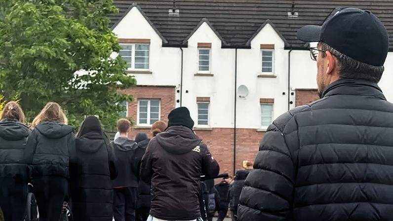 BBC People walk through a housing estate while a man watches from the roadside.