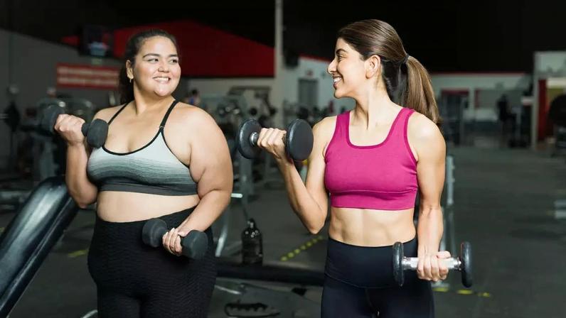 Duas mulheres com roupas de academia estão lado a lado levantando halteres. Elas estão sorrindo e aproveitando o treino. Uma delas está acima do peso, a outra tem peso considerado saudável e apresenta braços e abdômen visivelmente tonificados.