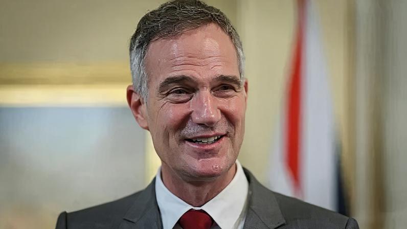 PA Media Peter smiles while standing in front of a blurry pale yellow background. He is wearing a light grey suit jacket, white shirt and deep red tie.