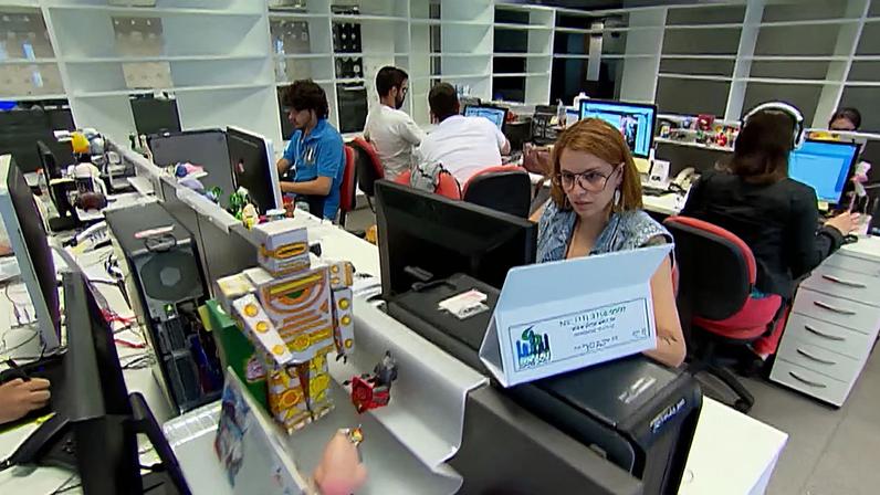 Um escritório eclético, sem cubículos e com as mesas decoradas, é registrado pela câmera. A expressão individual é bem-vinda no ambiente de trabalho.