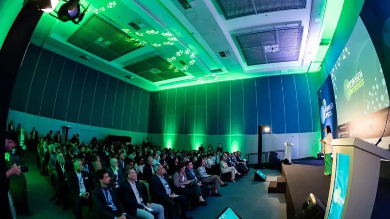 Auditório amplo com iluminação verde e azul, público atento em cadeiras escuras, e uma mulher no palco ao lado de um púlpito e telões exibindo Hydrogen Conferences
