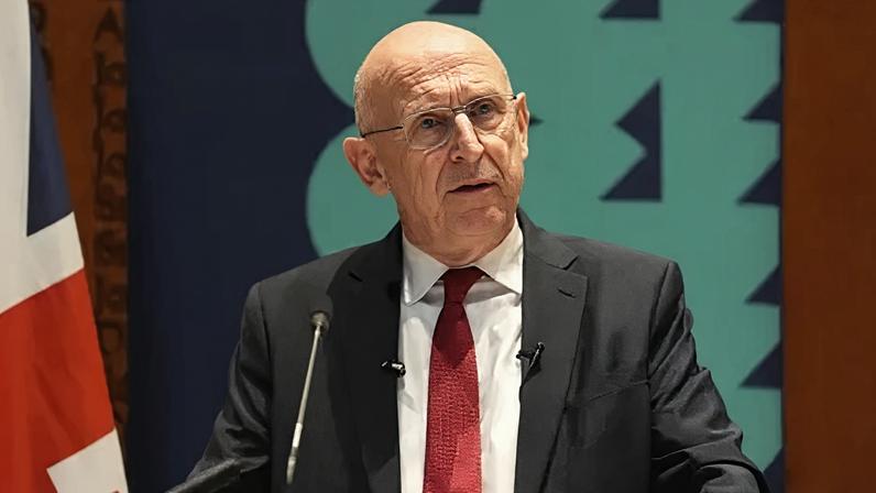 PA Media John Healey wearing a dark suit and red tie standing before a microphone