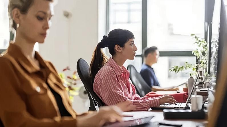 Getty Images A row of workers looking at their laptops in a brightly lit office. The icture focuses on a woman looking at a monitor in a red and white striped blouse, with her dark hair in a ponytail.