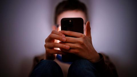 Getty Images A young person holding a smartphone in a dark room. We cannot see their face but it is illuminated by the phone's screen.