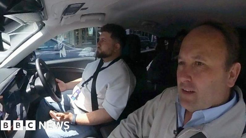 A safety driver is in the driver seat. His hands are off the wheel. A slightly concerned Transport Correspondent is in the passenger seat.