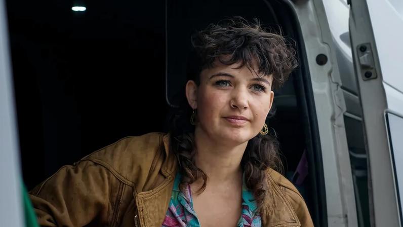 A young woman sits inside an open van holding a guitar.