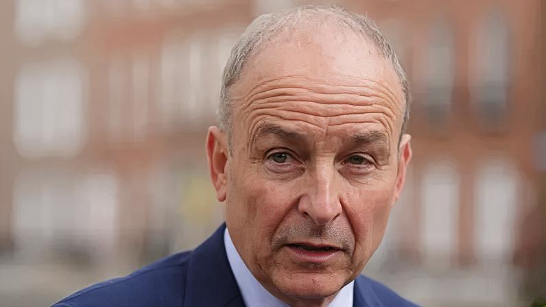 A man with thinning grey hair is standing outside in a suit. The background is out of focus.