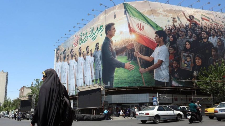 Uma mulher iraniana passa por um enorme outdoor em apoio à seleção nacional de futebol do Irã para a Copa do Mundo de 2026, instalado em um prédio na Praça Enghelab, em Teerã, em 18 de maio de 2026