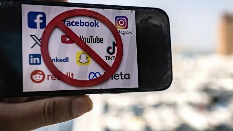 Mão segura um smartphone com tela exibindo logos de redes sociais como Facebook, YouTube, Instagram, X, LinkedIn, Snapchat, TikTok, Reddit e Meta, todos riscados por um círculo vermelho com barra diagonal, indicando proibição. O fundo está desfocado, com um céu claro e edifícios distantes