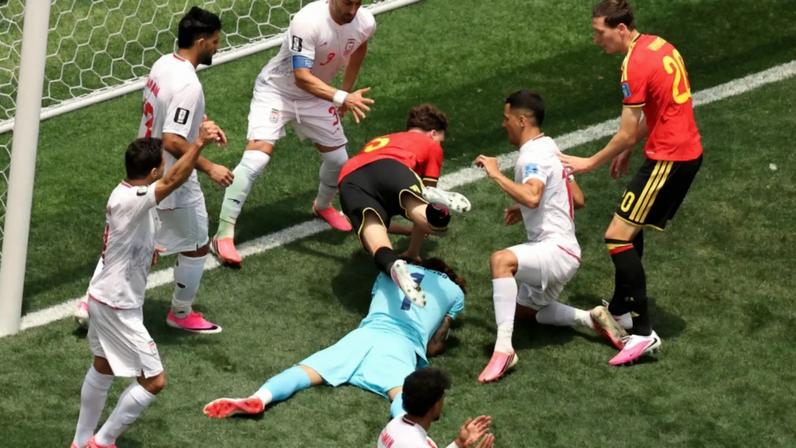Jogadores de futebol em campo verde, com um goleiro de uniforme azul claro deitado e um jogador de camisa vermelha e preta caindo sobre ele. Outros jogadores de branco e um de vermelho e preto observam a cena perto do gol.