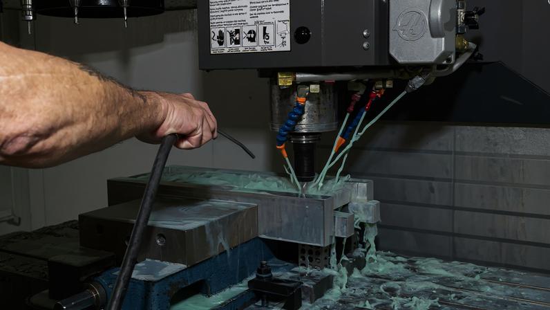 A technician is using a machine to drill pieces of silver metal; there is a light blue liquid coming out of jets next to the drill.