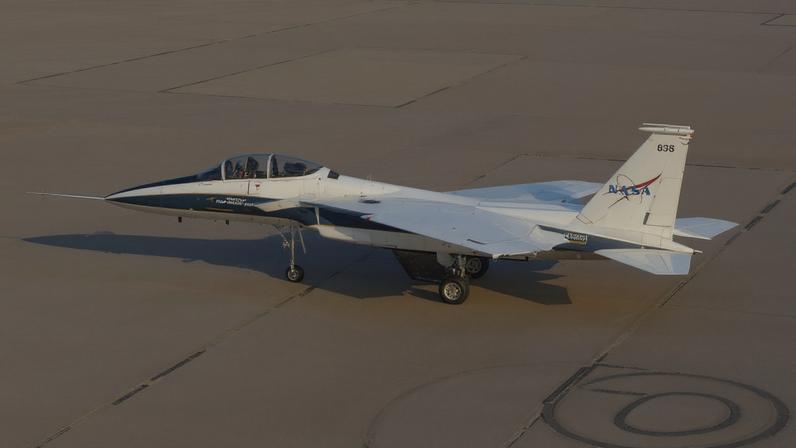 A white and blue painted jet aircraft flies above a tan lakebed with the gear and wheels out. A model wing hangs below the aircraft’s center line. The cockpit is closed and two pilots are visible inside with flight gear on.