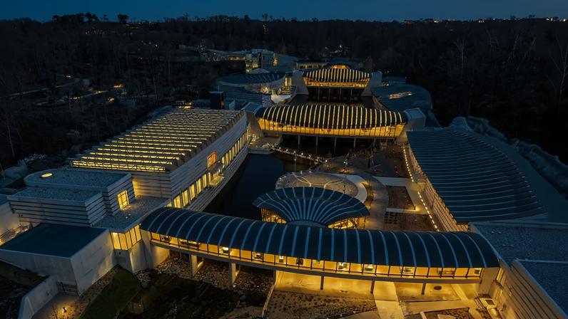 An aerial photo shows the Crystal Bridges Museum of American art at dusk, its galleries lit up from the interior, in a wooded landscape