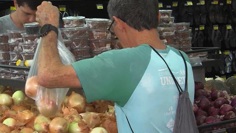 A imagem mostra um homem de costas selecionando cebolas em uma banca de hortifrúti dentro de um supermercado. Ele segura um saco plástico transparente com cebolas dentro, enquanto pega mais unidades da pilha exposta à frente. Ao redor, há outras bancas com ovos, produtos embalados e itens de mercearia nas prateleiras. Há também outra pessoa ao fundo manuseando embalagens.