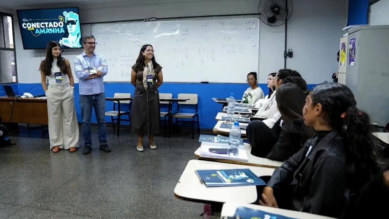 Três adultos, duas mulheres e um homem, sorriem em pé à frente de uma sala de aula com estudantes sentados. Um telão exibe Conectado Amanhã e uma imagem de óculos de realidade virtual.