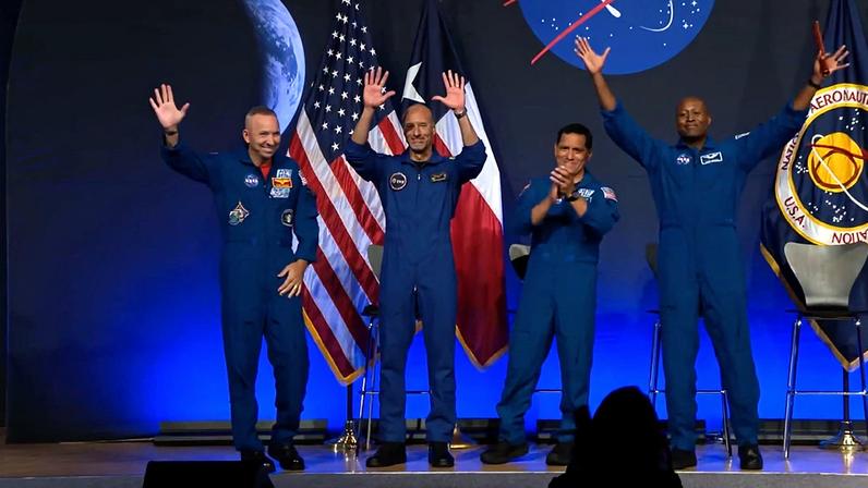 Quatro astronautas sorridentes em macacões azuis acenam para a plateia em um palco com bandeiras dos EUA e do Texas, e um logo da NASA