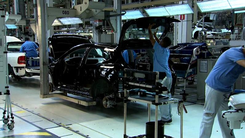 Em uma fábrica automotiva, três trabalhadores cumprem suas devidas funções e adicionam peças à carcaça de um carro.