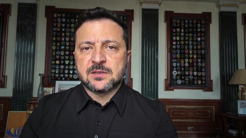 Volodymyr Zelensky, de camisa preta, fala diante da câmera em uma sala com quadros de distintivos nas paredes ao fundo