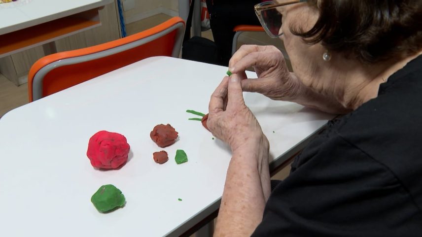 Pessoa idosa moldando massinha colorida sobre uma mesa, com pedaços em vermelho, verde e marrom ao redor