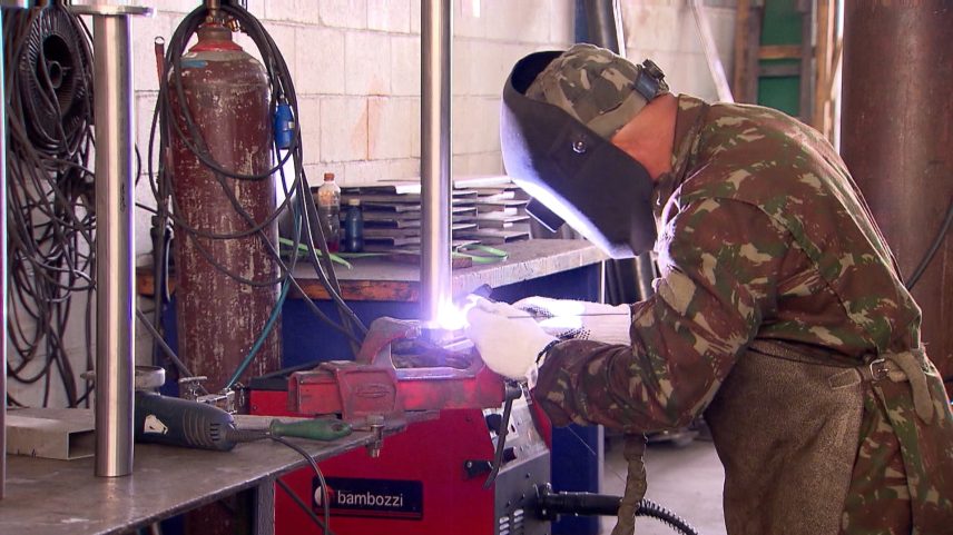 Pessoa realizando soldagem em oficina, usando máscara de proteção, luvas e uniforme camuflado com avental. O arco de luz indica o processo ativo de solda sobre peça metálica presa em objeto vermelho.