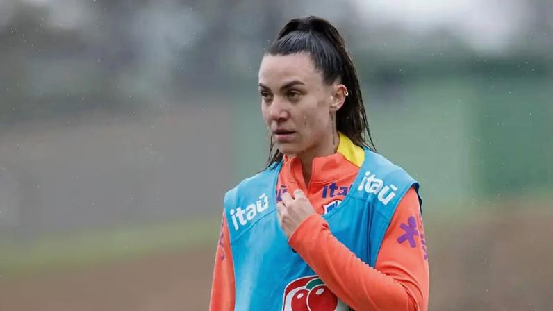 Jogadora de futebol durante um treino ao ar livre, vestindo uma camisa laranja de manga longa e um colete azul com logotipos de patrocinadores.