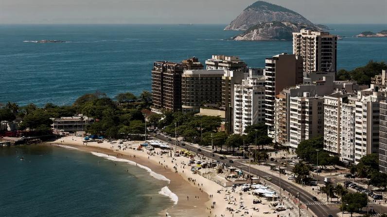 Vista aérea da Praia de Copacabana, Rio de Janeiro, com areia clara, mar azul-esverdeado e muitos banhistas. Prédios residenciais e comerciais margeiam a orla, com uma avenida movimentada.