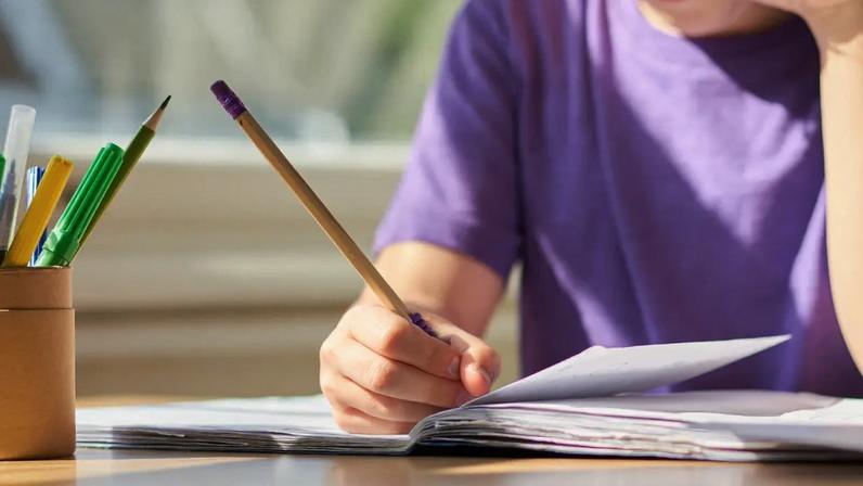 Criança de camiseta roxa escrevendo em um caderno aberto com um lápis, enquanto um porta-lápis com canetas e lápis coloridos está à esquerda, sobre uma mesa de madeira