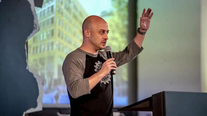 Homem careca, de perfil, com camiseta cinza e preta, segurando um microfone e gesticulando com a mão esquerda levantada, em um palco com projeção de prédio ao fundo