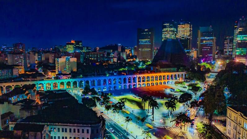 Vista aérea noturna dos Arcos da Lapa iluminados em azul e laranja, com a Catedral Metropolitana e arranha-céus ao fundo. Ruas movimentadas e palmeiras completam a paisagem urbana do Rio de Janeiro