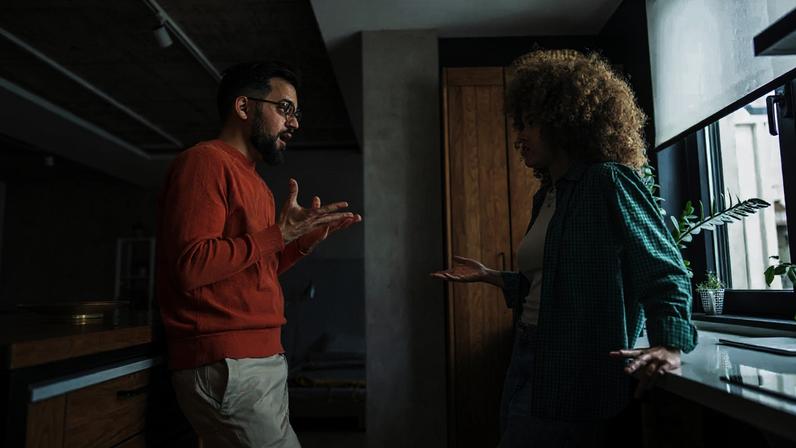Um homem e uma mulher, ambos com expressões sérias e gesticulando com as mãos abertas, discutem em uma cozinha moderna. Ele veste suéter laranja e calça clara, ela usa camisa xadrez verde e jeans.