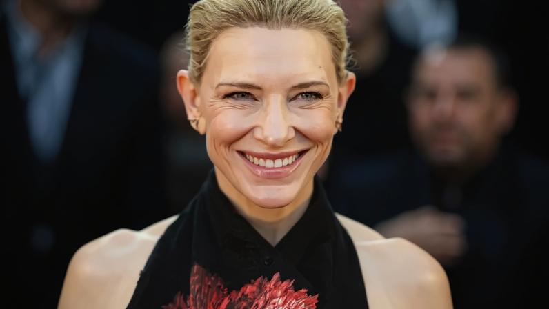 Cate Blanchett sorrindo, com cabelo loiro preso e brincos discretos, usando um vestido preto com uma grande flor vermelha bordada no centro do peito