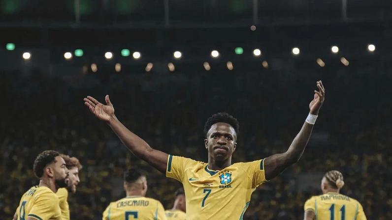 Jogador de futebol Vinicius Jr, de pele escura e cabelo curto, vestindo camisa amarela da seleção brasileira com o número 7, braços abertos e levantados, celebrando em campo com outros jogadores desfocados ao fundo, sob iluminação noturna do estádio